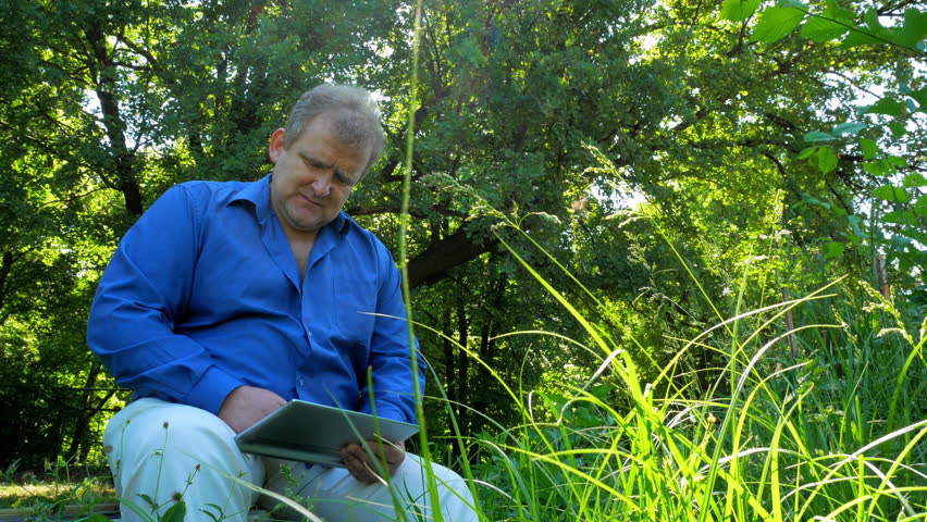 Adult man has a rest at the summer green park with tablet and reads news 