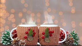 Closeup front view of two cups with hot drink on brown wooden table at holiday shiny lights background. White mugs  decorated with Christmas traditional elements. Real time full hd video footage.
