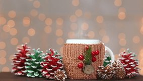 Closeup front view of one cup with hot drink on brown wooden table at holiday shiny Xmas lights background. White mug decorated with Christmas traditional elements. Real time full hd video footage.