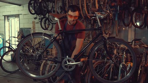 theme small business bike repair. young Caucasian brunette man wearing safety goggles, gloves and apron uses hand tool repairing and adjusting crank and pedal system bicycle in the garage workshop.: film stockowy