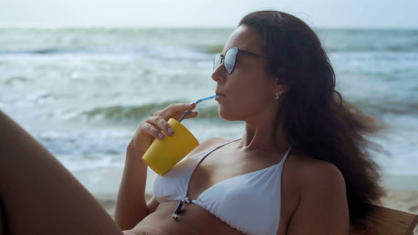 Close-up, A Beautiful Woman in a White Swimsuit and Sun Glasses Lies on a Wooden Deck Chair on the Sea Beach and Drinks a Cocktail