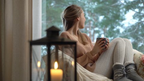 Cozy winter lifestyle. Happy young woman enjoy of cup of hot coffee sitting home by the big window with winter snow tree background. Self isolation, home quarantine. Stock Video