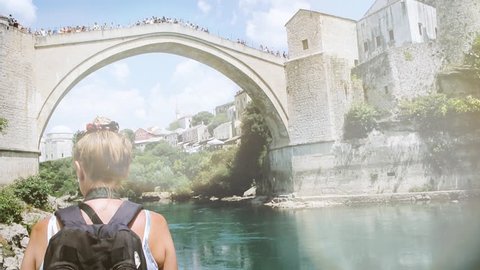 View of female travel blogger admiring and taking photos of UNESCO heritage Mostar Stari most bridge over picturesque Neretva river while other tourists are crossing it on a hot summer day