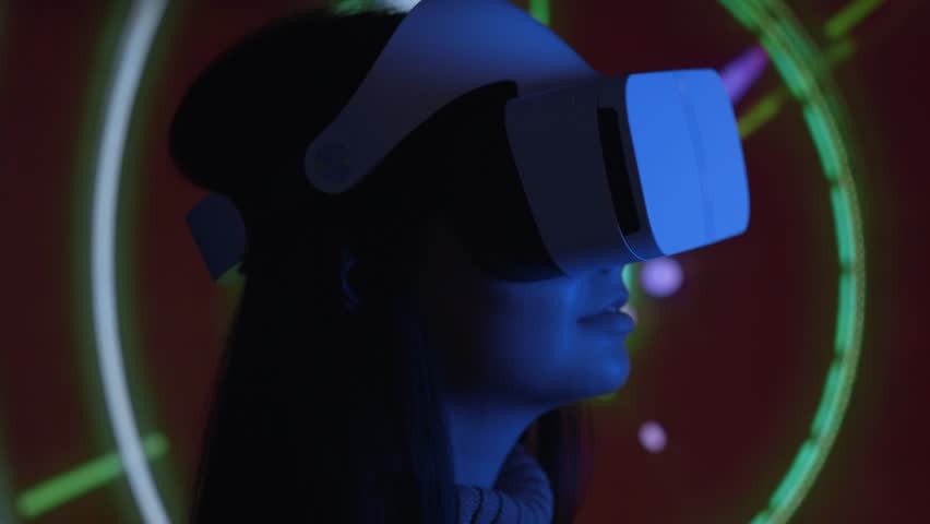 Cute young woman uses interactive VR headset, amaze and enjoys virtual reality. Wearing VR glasses or helmet to watch 360 video or play video game. Retro neon lighting, abstract background. Close up Royalty-Free Stock Footage #1016125636