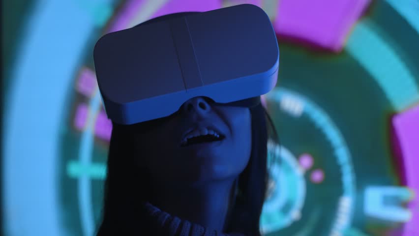 Cute young woman uses interactive VR headset, amaze and enjoys virtual reality. Wearing VR glasses or helmet to watch 360 video or play video game. Retro neon lighting, abstract background. Close up | Shutterstock HD Video #1016125636