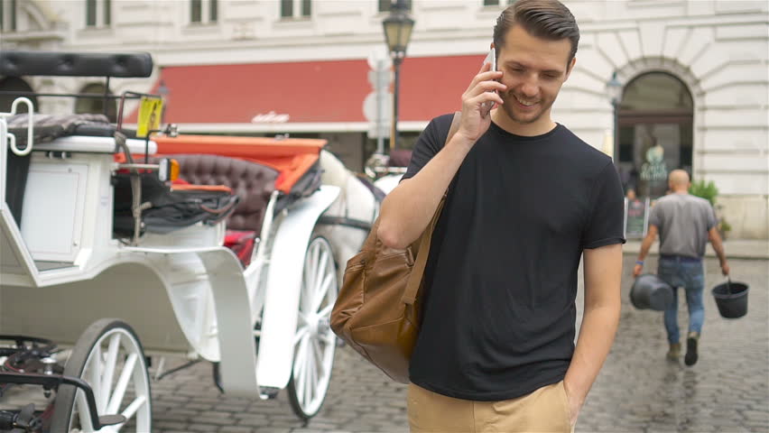 Tourist man enjoying a stroll through Vienna and talking by phone on background the beautiful horses in the carriage.