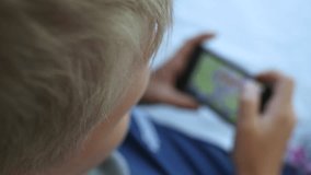 Top view closeup video of cute happy young kid of school age playing computer games using modern black smartphone. Focus at blond child's hair. Blurry device. Real time full hd footage.