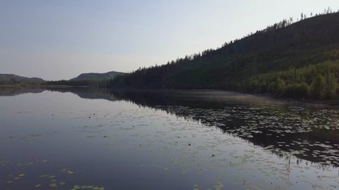 Chute Lake in morning light with water reflections mirroring forest, hill and sky. Fog or mist over surface. Drone aerial shot flying over water turning slowly.  BC Canada