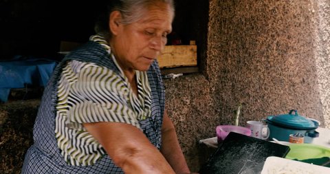 ZIRAHUEN, MEXICO - CIRCA MAY 2018 - Elderly senior Mexican woman rolling tortilla masa by hand and cooking them on a makeshift comal with a oil barrel lid over an open fire