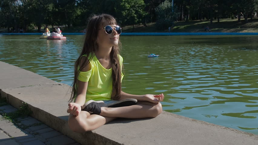 A child in a lotus pose in a city park. Happy little girl is meditating in the park by the water.