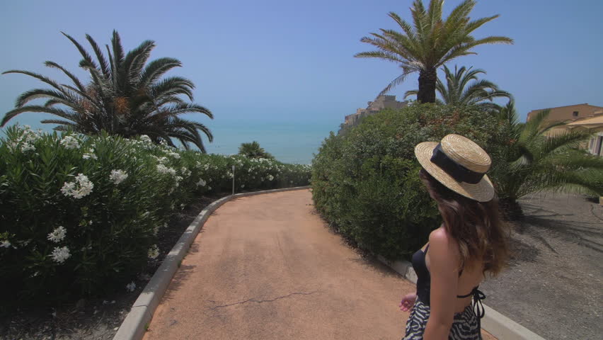 Happy young girl walking on hotel territory with very nice view on ocean.Brunette woman raising her hands and spinning. Dancing and turns around.Dressed in summer hipster clothes and hat. Slow motion 