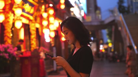 Young Chinese woman using mobile phone in street with traditional Chinese style building red lantern in the background in slow motion at Chengdu Sichuan China city