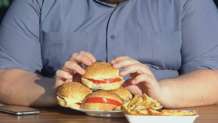 Eat fatty. Толстый человек с гамбургером. Жирный бургер и капсулы витаминов. Толстяк держит бургер Нижний ракурс. Eating fatty foods stock photo.