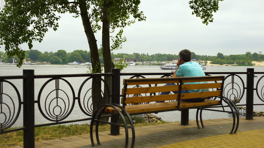 Male sitting on city bench enjoying river view in park, pleasure boat passing by