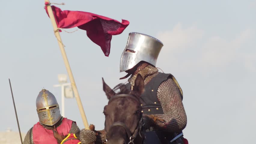 A man in armor and with a flag in his hand rides a horse past other men in historical costumes at a medieval tournament