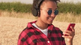 Slow motion tracking video clip of beautiful mixed race African American girl teenager young woman wearing blue sunglasses walking listening to music on wireless headphones and her smart phone