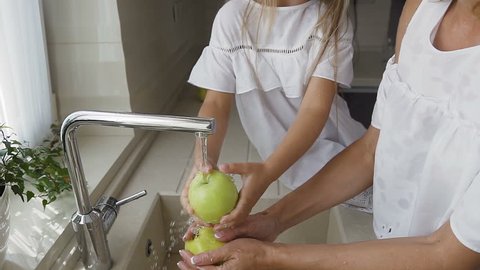 Young beautiful mom and her lovely two daughters together washes the green apples in the kitchen sink getting ready to cook fruits salad. One of the girls cuts an apple. They dressed in white clothes
