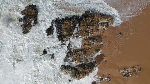 Aerial shot descending from sky down to white water waves crashing against sandstone rocks on sandy beach