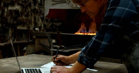 Blacksmith writing on clipboard while using laptop in workshop 4k
