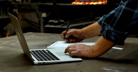 Close-up of Caucasian blacksmith writing on clipboard while using laptop in workshop 4k