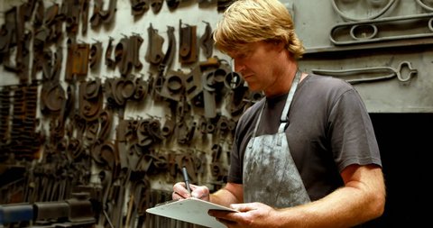 Attentive Caucasian blacksmith writing on clipboard in workshop 4k