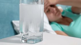 Closeup 4k video of glass of water with dissolving aspirin medicament on bedside table against woman suffering from headache lying in bed