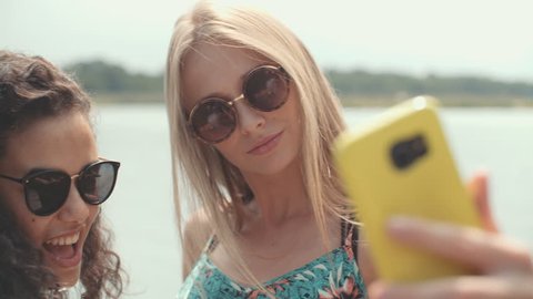 Two cheerful girls making selfies by a lake. Smiling two young women enjoying beautiful vacation. Video stock