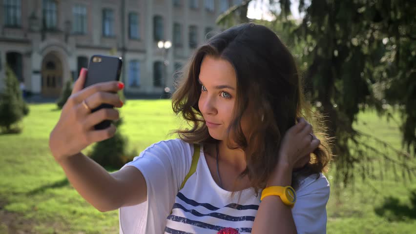 Portrait of a young smiling caucasian girl standing in the park and using a smartphone, making selfie, university in the background