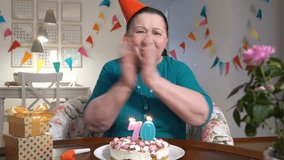 Happy senior woman holding cake to web camera while celebrating birthday with her family via video chat. Slow motion. Social distancing, self isolation.