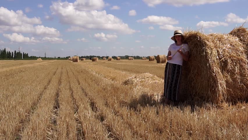 Calm young female in straw hat standing relaxed near huge bale of hay resting on hot summer day on sunny field landscape