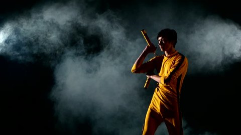 Costume Bruce Lee. the young man expertly twirling nunchaku.on black background. smoke, yellow clothing. Bruce Lee style.