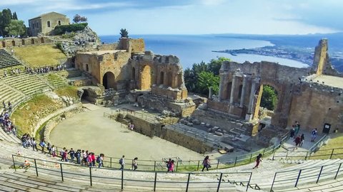 TAORMINA, ITALY - MAY 15, 2018: Ruins of ancient Greek theater in Taormina, famous resort in Sicily, Italy. Coast of Giardini-Naxos bay of Ionian sea and Mount Etna in smoke on background. Time Lapse