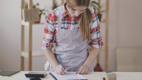 Small business. A girl in a striped apron and plaid shirt stands in a flower shop. The girl is engaged in calculation and planning, writes with a ballpoint pen and counting on the calculator. The girl