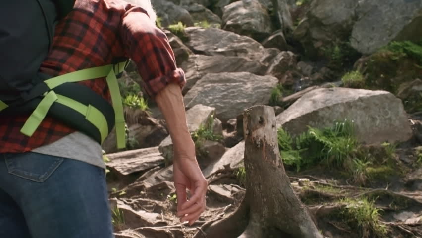Active senior couple with backpacks hiking in mountains: man holding hands with wife while climbing steep trail