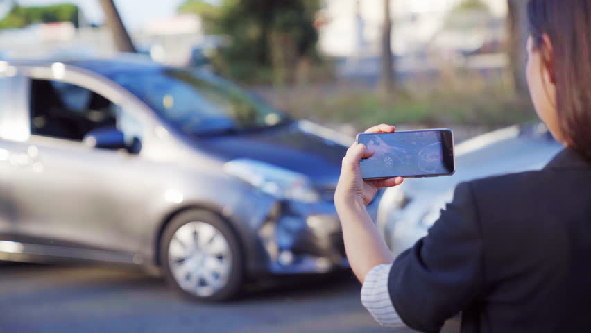 A female insurance agent taking photo of car accident on her smartphone. Two broken cars after car accident standing on the road. 4K UHD. Royalty-Free Stock Footage #1016291098