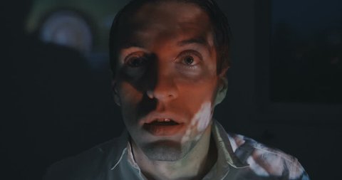 Close up of young man watching a horror video or film on TV or a computer monitor at home. He looks scared