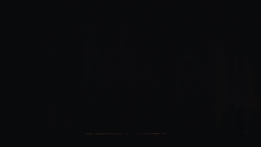 Young slim man opens two large massive doors and enters into darken empty hall leaving behind room with big chandalier consisting of several yellow bulb looking lamps mounted to ceiling.