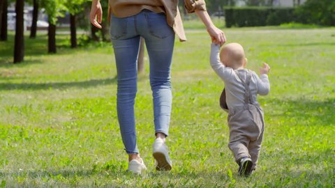 Tilt up shot of unrecognizable mother holding hand of baby boy in overalls and helping him walk on grass in park on summer day