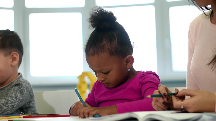 Diligent student. Cropped close up of a beautiful little African girl drawing at school smiling to the camera sitting next to her teacher education talent creativity ethnicity elementary classes kids  Royalty-Free Stock Footage #1016307118