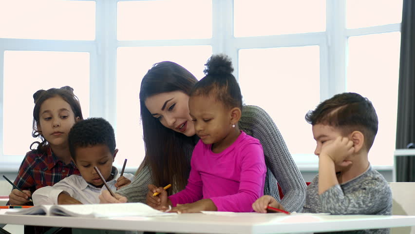 Helpful teacher. Cute little African girl sitting on the lap of her beautiful young female teacher and drawing during art class at elementary school education learning communication children ethnic Royalty-Free Stock Footage #1016307481