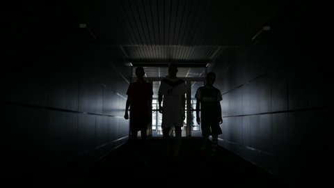 Three football players are walking along a dark tunnel to the football field.
