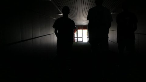 Three football players are walking along a dark tunnel to the football field. View from the back
