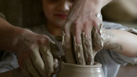 Tilt down with close up of smiling little girl and unrecognizable artisan throwing vase on spinning pottery wheel in ceramics class
