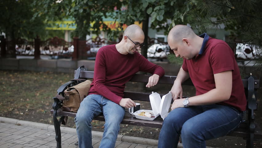 Men on a Park bench eating sandwiches with fries, drinking coffee from cups and talking