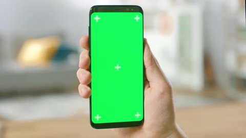 Close-up of a Man's Hand Holding Green Mock-up Screen Smartphone. Modern Mobile Phone. In the Background Cozy Living Room or Home Office. Shot on RED EPIC-W 8K Helium Cinema Camera.