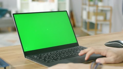 Man Uses Laptop with Green Mock-up Screen While Sitting at the Desk in His Cozy Living Room. Shot on RED EPIC-W 8K Helium Cinema Camera.