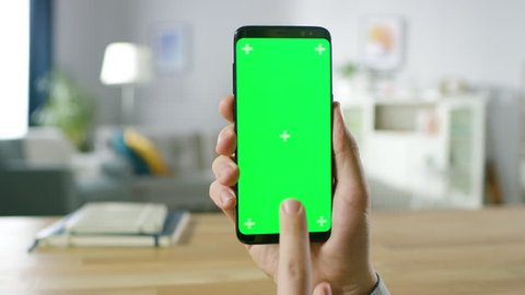Close-up of a Man Holding Green Mock-up Screen Smartphone and Using Touchscreen Gestures. Touching Mobile Phone Chroma Key Screen. In the Background Cozy Homely Atmosphere. Shot on RED EPIC-W 8K.