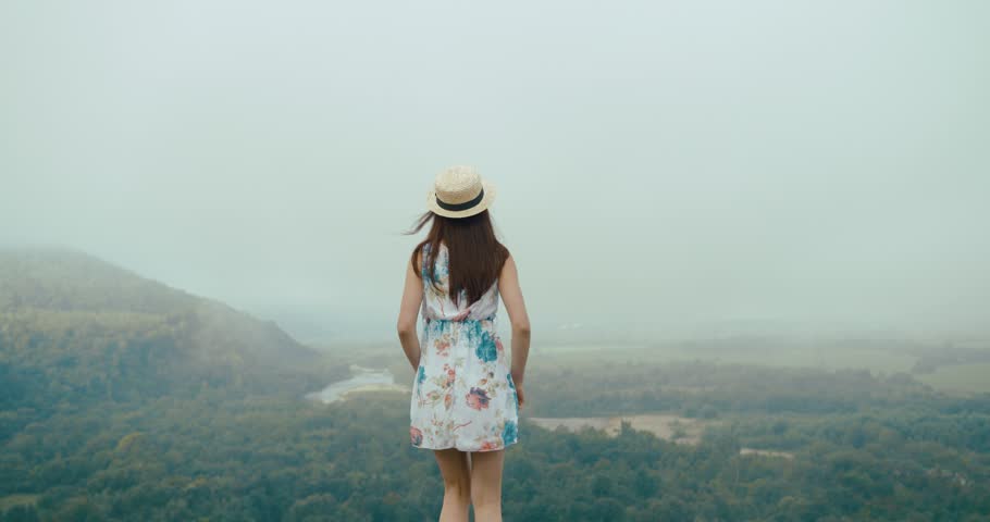 Charming young woman in a summer hat and dress stretches standing on the top of a mountain with great landscape behind her