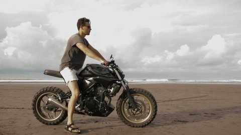 Young handsome hipster man sit on modern custom motorcycle cafe racer on the black sand ocean beach and drive away near the water. Adventure and travel concept. Surfing spot with ocean waves.
