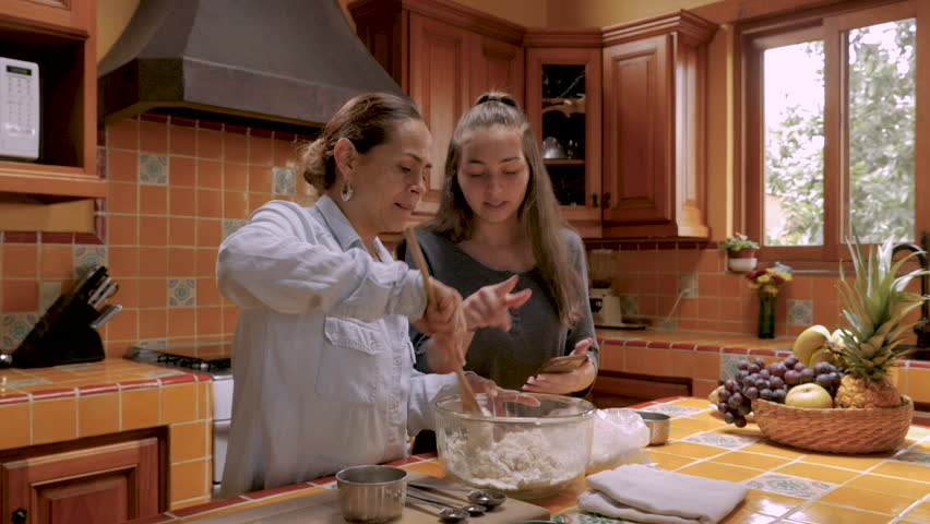 Happy smiling teenager girl referencing an app on her cell phone to help her Mexican mother bake in their kitchen together - slow motion push in Royalty-Free Stock Footage #1016337832